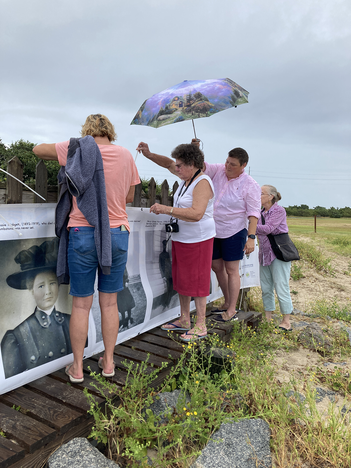 Jean Hooper, center, whose husband, grandparents and great-grandparents are buried at the Salvo Community Cemetery, helps secure the banner to the fence, as Jenny Creech, who also has kin buried here, shields her from drizzle. Photo: Justin Cook 