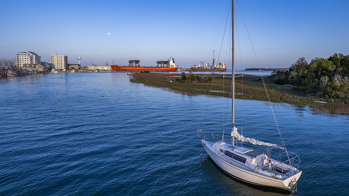 A sailboat is anchored in the cut near Sugarloaf Island along the Morehead City waterfront in Carteret County, with two cargo ships shown berthed at the North Carolina Port of Morehead City in the background. Photo: Dylan Ray