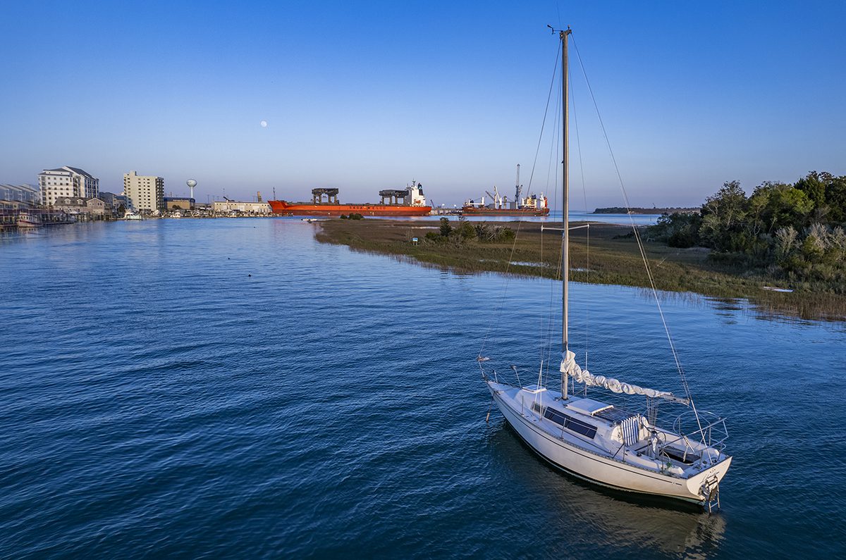 A sailboat is anchored in the cut near Sugarloaf Island along the Morehead City waterfront in Carteret County, with two cargo ships shown berthed at the North Carolina Port of Morehead City in the background. Photo: Dylan Ray