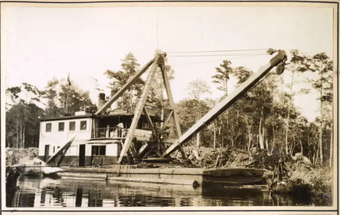 This is the U.S. Army Corps of Engineers’ derrick barge No. 14 at work on Turner’s Cut,  a canal in Camden County, N.C., September 1931. Turner’s Cut was originally dug in the 1850s, presumably by slave laborers (at least if its construction was like that of all the state’s other antebellum ship canals). The 4.4-mile-long canal was designed so that vessels headed north to, or leaving south from, the Dismal Swamp Canal would no longer have to traverse Joyce’s Creek, a shallow, narrow, and winding stream that flows into the Pasquotank River and which was often called the “Moccasin Track.” The canal ran from South Mills, N.C., at the southern end of the Dismal Swamp Canal, to a broader, more navigable point on the Pasquotank closer to Elizabeth City. In 1929, the Corps of Engineers had purchased the Dismal Swamp Canal  and made that whole route part of the Intracoastal Waterway (IWW), the grand network of canals and natural waterways that the Army Corps of Engineers was still in the process of building at that time. At the time of this photograph, the Corps of Engineers had completed the IWW from the Delaware River to Beaufort, N.C.. Another section of the IWW, extending from Beaufort to Wilmington, was under construction. In this photograph, the No. 14’s crew is removing a 2,500-foot-long shoal that had built up just below the canal lock at South Mills.  Source: Office of History, HQ, U.S. Army Corps of Engineers
