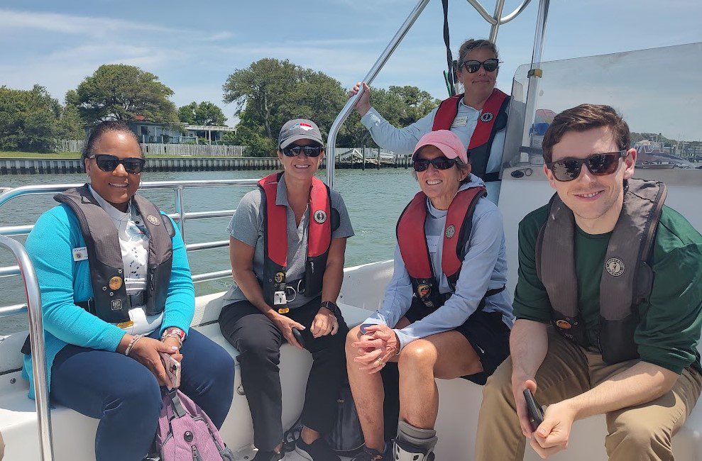 Seated, from left, Beaufort Mayor Sharon Harker, Rep. Celeste C. Cairns, R-Carteret and Craven counties, Rep. Pricey Harrison, D-Guilford, and Skyler Golann, NCDEQ legislation liaison, and standing, Central Sites Manager Paula Gillikin travel by boat Friday along Taylor’s Creek. Photo: Jennifer Allen