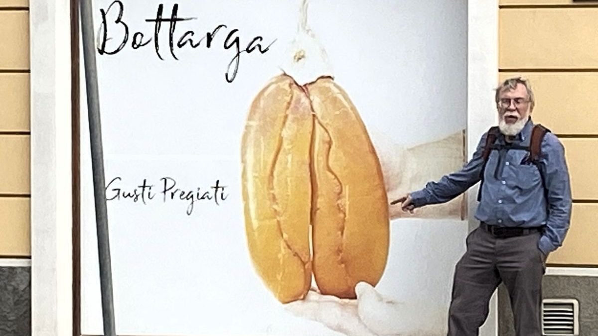 Author points to a giant poster of dried mullet roe (bottarga di muggine) in front of a store specializing in bottarga, smoked mullet, and other local seafood products, in Cabras, Sardinia, arguably the “Mullet Capital of the World.” Photo: Lida Pigott Burney